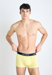 Gele boxershorts met een zwarte Lacoste tailleband, nauwsluitend ontwerp, gladde textuur en middellange benen. Model past de tailleband aan.