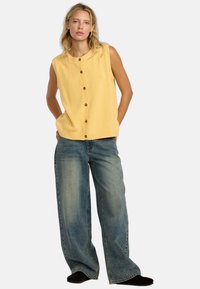 Gilet sans manches en tricot jaune avec des bords côtelés, présentant une fermeture à boutons sur toute la longueur à l'avant. Associé à un jean en denim bleu ample.