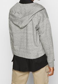 Personne portant une veste à capuche grise sur un t-shirt noir et un pantalon cargo beige, vue de dos sur un fond uni.