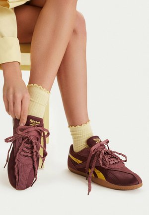 Una persona con calzini gialli e sneakers Reebok bordeaux è seduta e si sta sistemando una scarpa su uno sfondo bianco.