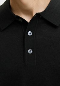 Juoda megztinė polo su apykakle, dviem pilkomis mygtukais ir tekstūruotu audiniu. Artimas vaizdas į viršutinę drabužio dalį.