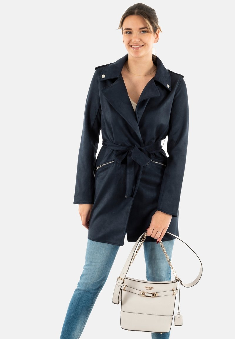 Trench-coat bleu marine en matériau doux, avec une taille ceinturée, de larges revers et deux poches zippées. Sac à main blanc avec une bandoulière en chaîne.