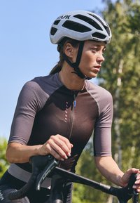 Short-sleeved cycling jersey in dark brown and gray, featuring a front zipper and pink logo, worn with a white helmet on a bike.