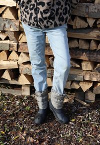 Schwarze wasserdichte Stiefel mit gepolstertem grauem Obermaterial und verstellbarem Schaft, getragen zu hellblauen Jeans, vor einem Hintergrund aus Holzstämmen.