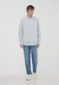 Camicia azzurra a righe bianche con maniche lunghe, abbinata a jeans blu medio e sneakers bianche. Colletto semplice, vestibilità dritta.