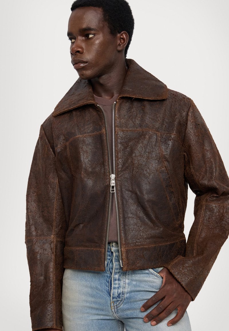 Brown leather jacket with a wide collar, full zip front, and textured surface. Features include visible stitching and side pockets.