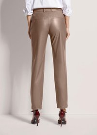 Vrouw in taupe straight-leg broek en bordeauxrode hoge hakken, staand tegen een neutrale achtergrond, gezien van achteren.