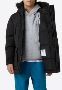 Μαύρο μπουφάν με επένδυσ η, με κουκούλα με γούνα, φερμουάρ, κλείσιμο με velcro και λογότυπο. Φορώντας από πάνω γκρι μπλούζα με κουκούλα και μπλε παντελόνια.