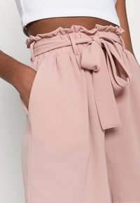 Gros plan sur une personne portant un short taille haute texturé rose clair avec une ceinture en tissu nouée en nœud et une main dans la poche.