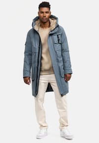 Kék bélelt parká, bolyhos kapucnival, elülső zsebekkel és cipzárral. Bézs pulóver és világos bézs nadrág fölött viselve, fehér sportcipővel párosítva.