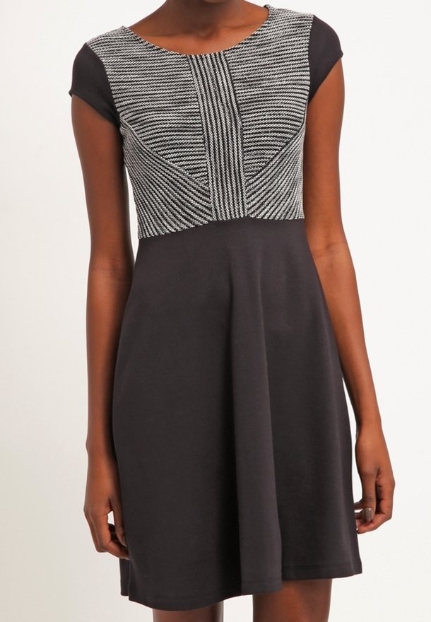 Femme portant une robe noire à manches courtes avec un corsage cintré, texturé, à motifs gris et noirs, et une jupe évasée.