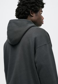 Σκούρα γκρι φούτερ με μεγάλη, μαλακή κουκούλα. Διαθέτει χαμηλωμένους ώμους και άνετη εφαρμογή. Ομαλή υφή με ορατές ραφές.