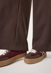 Brūni platajiem biksēm ar gludu tekstūru, kas apvienotas ar bordo un pelēkām kedām, kurām ir teksturēta virsma un baltas šņores.