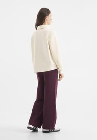 Kremowa bluzka z długim rękawem i kołnierzykiem, zestawiona z luźnymi, ciemnofioletowymi spodniami. Modelka stoi tyłem do kamery, mając na sobie białe i czarne sneakersy.