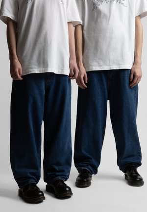 To personer iført hvide t-shirts med grafisk tryk, løstsiddende mørkeblå jeans og sorte lædersko, stående side om side mod en ensfarvet baggrund.