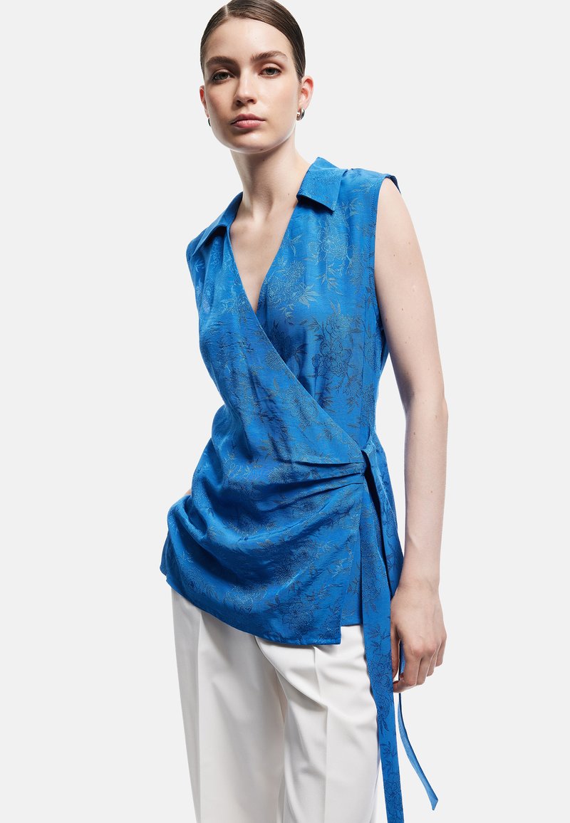 Femme portant un haut cache-cœur bleu fleuri sans manches avec col, associé à un pantalon blanc, debout devant un fond uni clair.