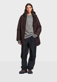 Femme portant une veste d'hiver marron foncé, un pull gris, un pantalon noir et des chaussures de randonnée, debout devant un fond clair uni.