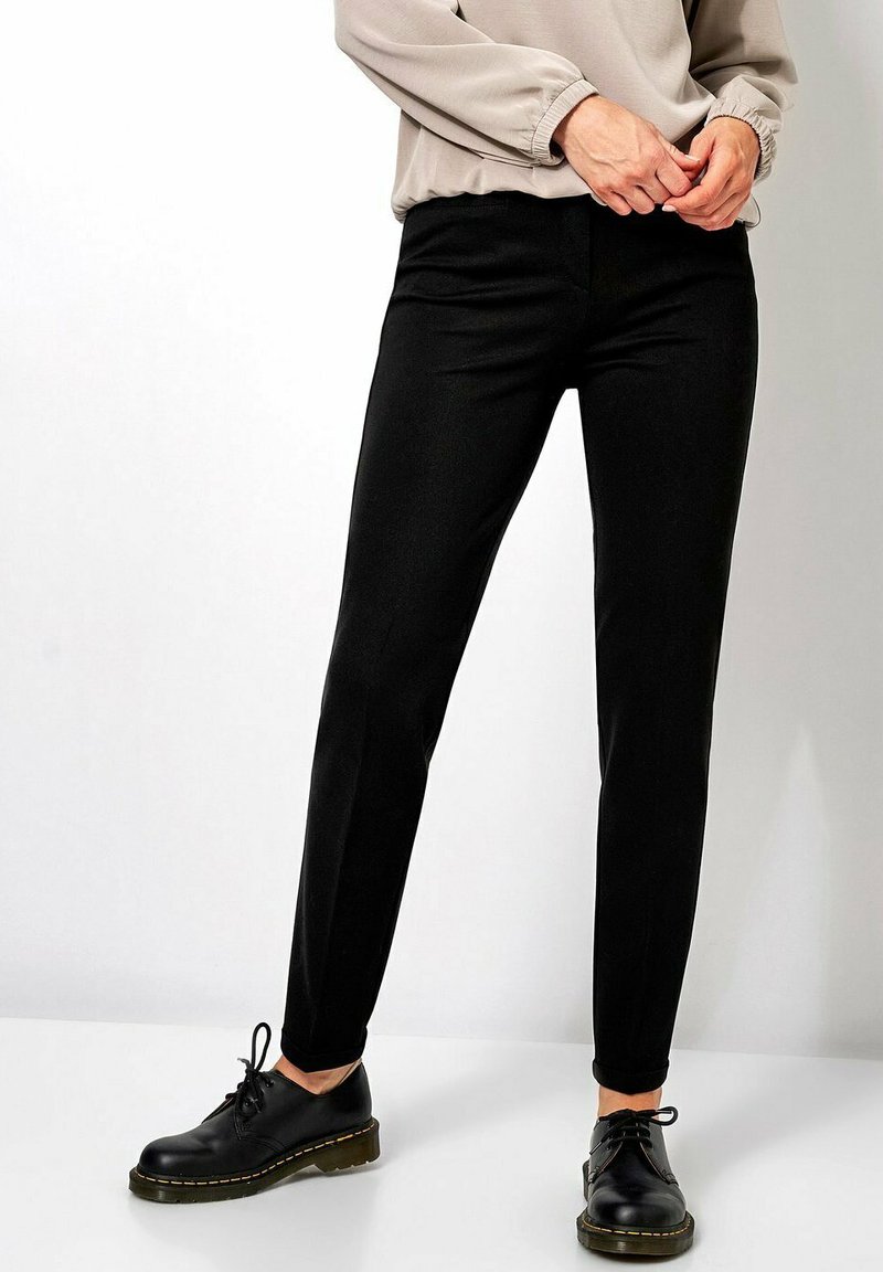 Zwarte slim-fit broek met een gladde textuur, voorzien van een middelhoge taille en spits toelopende pijpen, gecombineerd met zwarte veterschoenen.