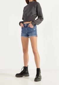 Grauer, verkürzter Sweatshirt mit gerafftem Saum, kombiniert mit blauen Denim-Shorts mit umgeschlagenen Säumen und Abnutzungsspuren, ergänzt durch schwarze Kampfstiefel.
