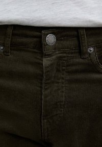 Pantalones de pana verde oscuro con una tela texturada, botones de metal y una marca en la cinturilla. Costuras visibles y bolsillos.