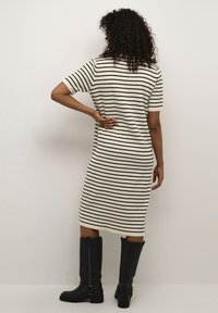 Robe en maille rayée crème et noire, longueur genou avec manches courtes. Portée avec de grandes bottes noires ornées de boucles.