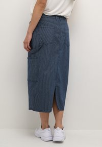 Falda midi de tela vaquera con un patrón de rayas verticales, presenta una abertura en la parte posterior, bolsillos y una cinturilla ajustada, combinada con zapatillas blancas.