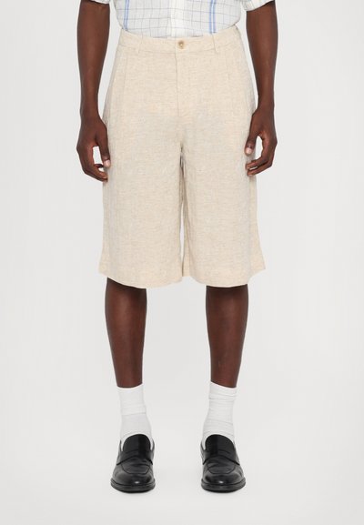 Pantalones cortos de beige claro con una tela texturizada, que cuentan con un cierre de botón y pinzas. Combinados con calcetines blancos y zapatos negros.