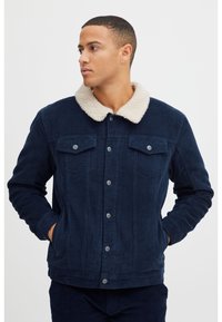 Chaqueta de pana azul marino con cuello de piel sintética crema, cierre frontal de botones, dos bolsillos en el pecho y bolsillos laterales para las manos. Detalle de tejido texturizado.