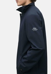 Chaqueta ecológica azul marino con cuello alto, bolsillos laterales y detalles de botones. Presenta un logo en la manga.