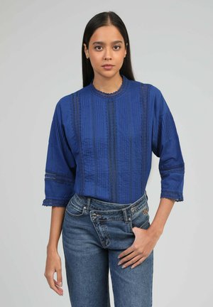 Femme aux longs cheveux bruns portant un chemisier bleu texturé à manches trois-quarts et un jean taille haute bleu dépareillé.