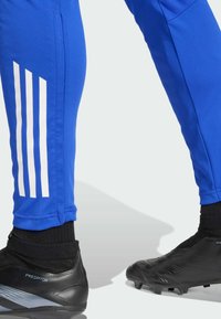 Blauwe sportbroek van gladde stof met drie witte strepen langs de zijkant. Zwarte voetbalschoenen met een gestructureerd oppervlak en enkelondersteuning.