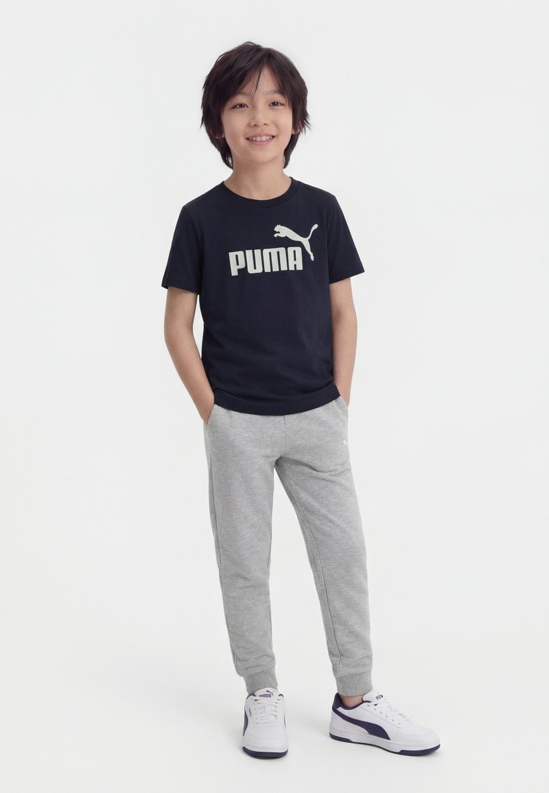 Jonge jongen staand met handen in de zakken, draagt een zwarte Puma T-shirt, grijze joggingbroek en witte Puma sneakers, tegen een witte achtergrond.