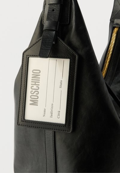 Κοντινή λήψη μαύρης δερμάτινης τσάντας με ραμμένη θήκη για ετικέτα που εμφανίζει κενή ετικέτα Moschino για όνομα και διεύθυνση.