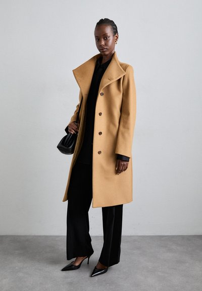 Camel wool coat with a wide collar, button closures, and side pockets; paired with black trousers and pointed black heels.