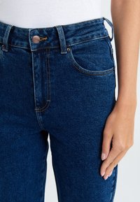 Džiny s vysokým pasem v tmavě modré barvě s měděným knoflíkem, designem s pěti kapsami a kontrastním prošíváním, s měkkým, texturovaným povrchem.