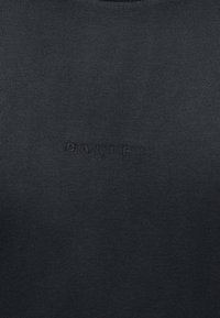 Μαύρο ύφασμα με διακριτικό, ανάγλυφο λογότυπο Oakley στο κέντρο της επιφάνειας, που παρουσιάζει λεπτομέρεια μινιμαλιστικού υφασμάτινου σχεδίου με το εμπορικό σήμα.