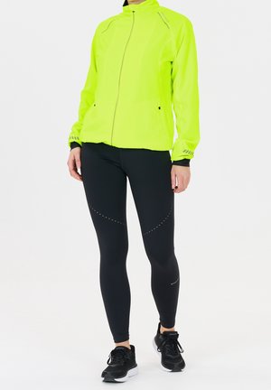 Persona che indossa una giacca zippata giallo neon, leggings neri con dettagli riflettenti a puntini e scarpe da corsa nere, in piedi su uno sfondo bianco.