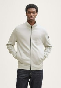 Veste zippée gris clair en tissu lisse, dotée d'un col haut, d'une fermeture éclair noire et d'une texture subtile. Assortie à un jean foncé.