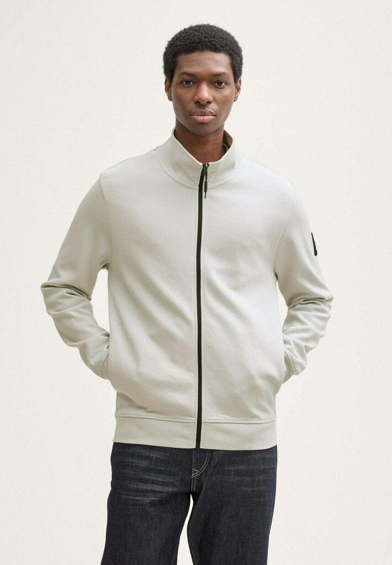 Veste zippée gris clair en tissu lisse, dotée d'un col haut, d'une fermeture éclair noire et d'une texture subtile. Assortie à un jean foncé.