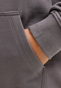 Una mano parzialmente dentro della tasca anteriore di un felpa grigia e texturizzata, con cuciture visibili e polsini a costine.