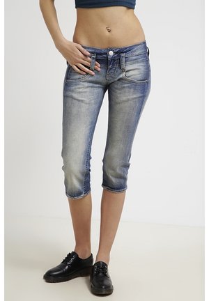 Femme portant un pantalon capri en denim bleu délavé, des chaussures décontractées noires et un haut court bleu marine, debout avec une main sur la hanche.