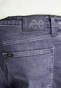 Temno sivi denim kavbojke z usnjeno nalepko, na kateri je napisano "Lee." Vidna je šivana obdelava zadnjega žepa, kar prikazuje klasičen dizajn.
