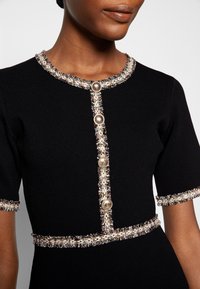 Haut noir à manches courtes avec bordure en tweed décorative et quatre boutons dorés sur le devant, porté par une personne à la carnation moyenne foncée.