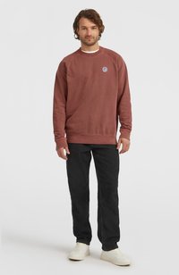 Donkerrode sweatshirt met lange mouwen en een ronde hals, voorzien van een klein cirkelvormig logo. Gecombineerd met zwarte broek en witte sneakers.