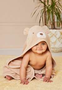 Beige bear hooded towel features ears and an embroidered face, enveloping a seated baby on a textured yellow rug. Background plant in a pot.
