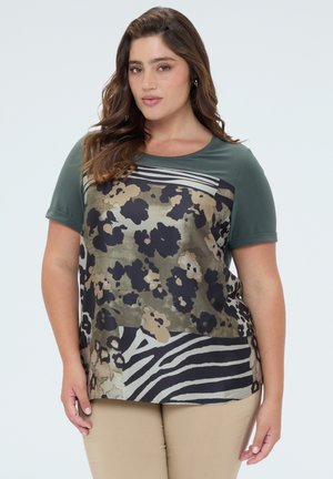 Kvinde med langt brunt hår iklædt en grøn og mønstret bluse med blomster- og zebra-print og beige bukser på ensfarvet baggrund.