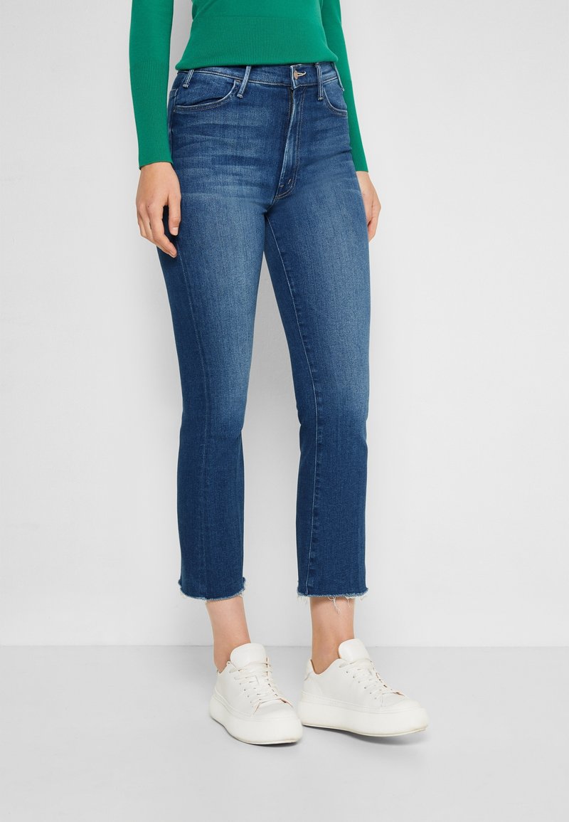 Höga, korta blå jeans med fransad nederkant, tillverkade av denimtyg. Bärs med vita sneakers och en grön långärmad topp.