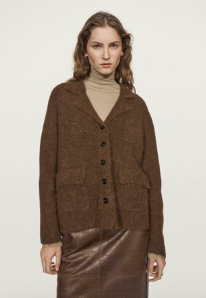 Woman with wavy hair wearing a beige turtleneck, brown fuzzy buttoned cardigan, and dark brown textured skirt against a plain background.