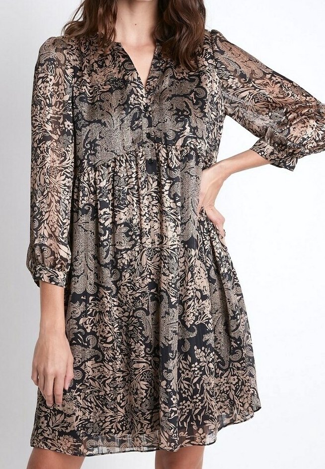 Femme portant une robe longue jusqu'aux genoux à manches longues avec des motifs floraux et cachemire noirs et beiges, une main sur la hanche et l'autre pendant le long du corps.