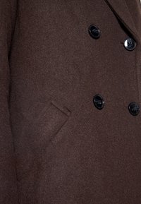 Hnědý vlněný dvouřadý kabát s dvěma velkými černými knoflíky a bočním kapsou s texturovaným povrchem.